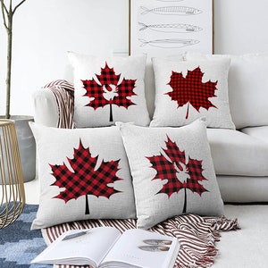Puede incluir: Cuatro almohadas blancas con diseños de hojas de arce de cuadros rojos y negros. Las almohadas están dispuestas en un sofá blanco con una manta de rayas rojas y blancas.