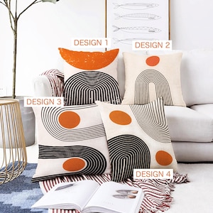 May include: Four decorative cushions with abstract designs in orange, black, and white are displayed on a white sofa. The cushions feature geometric shapes, including arches and circles. The cushions are labelled "DESIGN 1", "DESIGN 2", "DESIGN 3", and "DESIGN 4".
