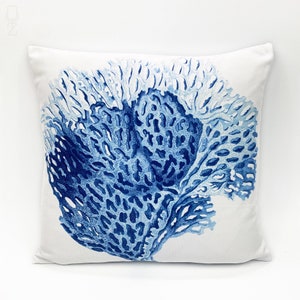 Op de afbeelding: Een wit kussen met een blauw koraalontwerp. Het koraal is lichtblauw en donkerblauw en bevindt zich in het midden van het kussen.