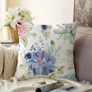 Könnte beinhalten: Ein weißes Kissen mit einem Blumenmuster mit rosa Rosen, weißen Anemonen und blauen Sukkulenten. Das Kissen hat ein aquarellartiges Design und ist perfekt, um Ihrem Zuhause einen Hauch von Natur zu verleihen.