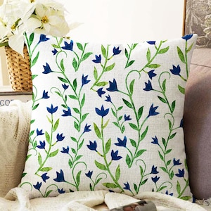 May include: A white throw pillow with a blue and green floral pattern. The pillow features a repeating pattern of blue bell-shaped flowers and green leaves on a white background.