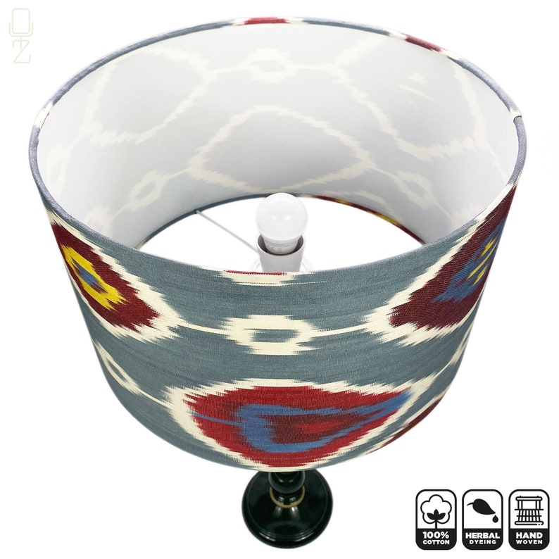 May include: A gray and white lampshade with a red, blue, and yellow ikat pattern. The lampshade is made of 100% cotton and is hand-woven. The lampshade is sitting on a black lamp base.