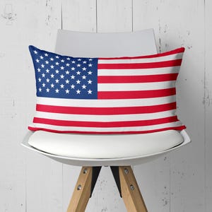 Può includere: Cuscino rettangolare con il design della bandiera americana. Presenta un cantone blu con stelle bianche e strisce rosse e bianche. Il cuscino è su una sedia bianca con gambe in legno, contro un muro di legno bianco.