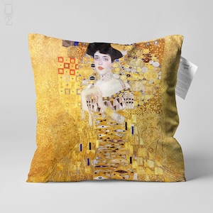May include: A square throw pillow with a gold background featuring a portrait of a woman with dark hair and a gold necklace. The woman is wearing a patterned dress with gold accents. The background is filled with geometric shapes and swirls in gold.
