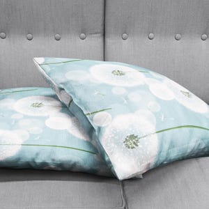 K&ouml;nnte beinhalten: Zwei hellblaue Kissenbez&uuml;ge mit einem wei&szlig;en L&ouml;wenzahnmuster. Die Kissenbez&uuml;ge liegen auf einem grauen Sofa.