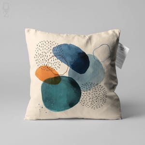 May include: A beige throw pillow with a geometric pattern of blue, teal, and orange watercolor shapes. The shapes are outlined in black and have a speckled pattern.