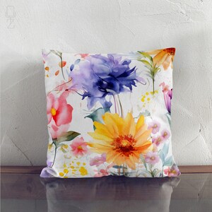 May include: Square throw pillow with a watercolor floral design. The pillow features a white background with colorful flowers in shades of yellow, purple, pink, and orange. The flowers are painted in a loose, artistic style.