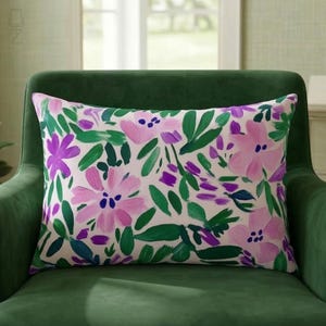 May include: Rectangular decorative pillow with a floral pattern. The pillow features large pink and purple flowers with green leaves on a white background. The pillow is placed on a green armchair.