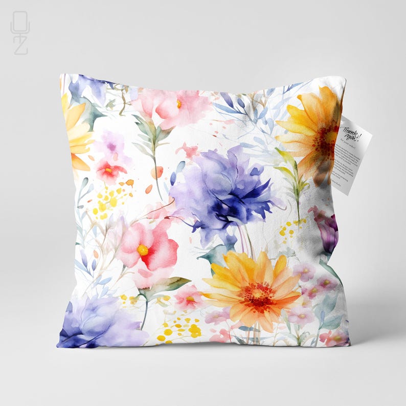 May include: A square pillow with a watercolor floral pattern. The design features a variety of flowers in shades of pink, yellow, orange, and purple, set against a white background. The pillow has a tag attached with text.