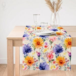 May include: A floral table runner featuring a watercolor design with orange gerbera daisies, purple cornflowers, and pink roses. The runner is set on a light wood table, with glassware and a vase of dried flowers in the background.