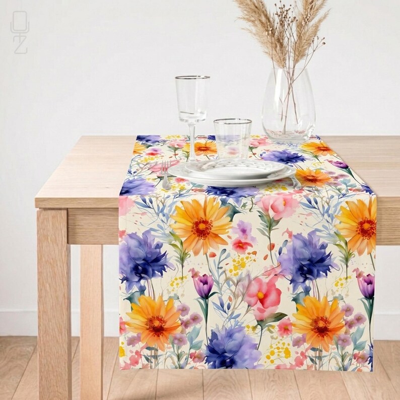 May include: A floral table runner featuring a watercolor design with orange gerbera daisies, purple cornflowers, and pink roses. The runner is set on a light wood table, with glassware and a vase of dried flowers in the background.
