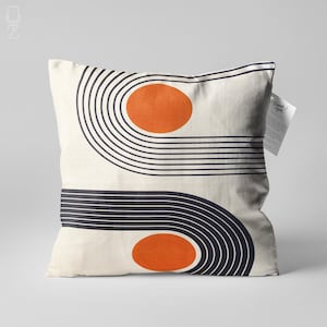 Op de afbeelding: Vierkant kussen met een crèmekleurige achtergrond. Het heeft een geometrisch ontwerp met twee oranje cirkels en gebogen zwarte lijnen. Een label met tekst is aan de zijkant bevestigd. Een decoratief woonaccessoire.