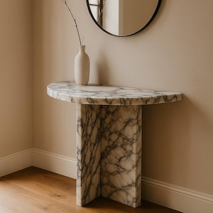 Puede incluir: Una consola de mármol con una tapa semicircular y una base geométrica única. La mesa presenta un patrón veteado blanco y gris. Un pequeño jarrón beige con una ramita está sobre la mesa, y un espejo redondo negro cuelga de la pared.