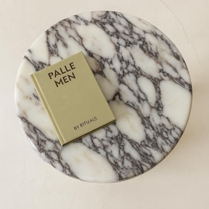 May include: A round marble tray with a white and gray veined pattern. A small, olive-green book titled "PALLE MEN" by Rituals rests on the tray. The tray is on a white surface.