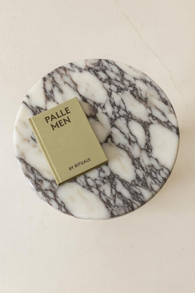 May include: A round marble tray with a white and gray veined pattern. A small, olive-green book titled "PALLE MEN" by Rituals rests on the tray. The tray is on a white surface.