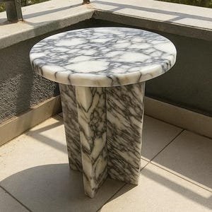 Op de afbeelding: Een ronde marmeren bijzettafel met een wit en grijs patroon. De tafel heeft een cirkelvormig blad en een voetstuk met drie steunpoten. De tafel is ongeveer 50 cm in diameter en 50 cm hoog.