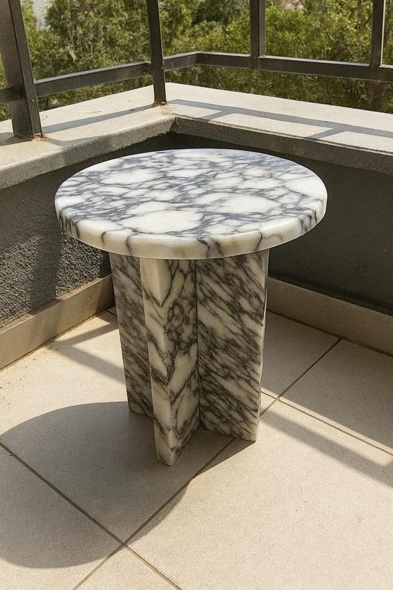 May include: A round marble side table with a white and gray pattern. The table has a circular top and a pedestal base with three supporting legs. The table is made of marble and is suitable for indoor or outdoor use.