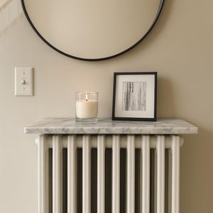 Puede incluir: Una vela encendida en un vaso transparente sobre una estantería de mármol encima de un radiador blanco. También se ven una impresión en blanco y negro enmarcada y un espejo circular negro. La pared está pintada de color beige neutro.
