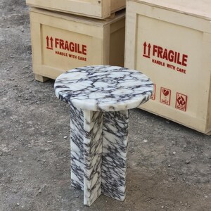 May include: A small, round marble side table with a white and gray veined pattern. The table has a circular top and a pedestal base. The background includes wooden crates with "FRAGILE" labels.