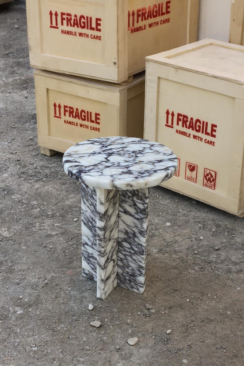 May include: A small, round marble side table with a white and gray veined pattern. The table has a circular top and a pedestal base. The background includes wooden crates with "FRAGILE" labels.