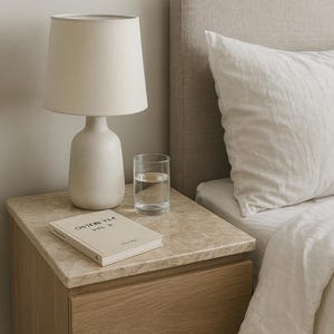 Könnte beinhalten: Ein Schlafzimmer mit einem Nachttisch mit einer Lampe, einem Glas Wasser und einem Buch. Die Lampe hat einen cremefarbenen Schirm und Sockel. Das Buch trägt den Titel "OSTERS 334 VOL. 2". Im Hintergrund ein Bett mit weißem Kissen und Bettdecke.