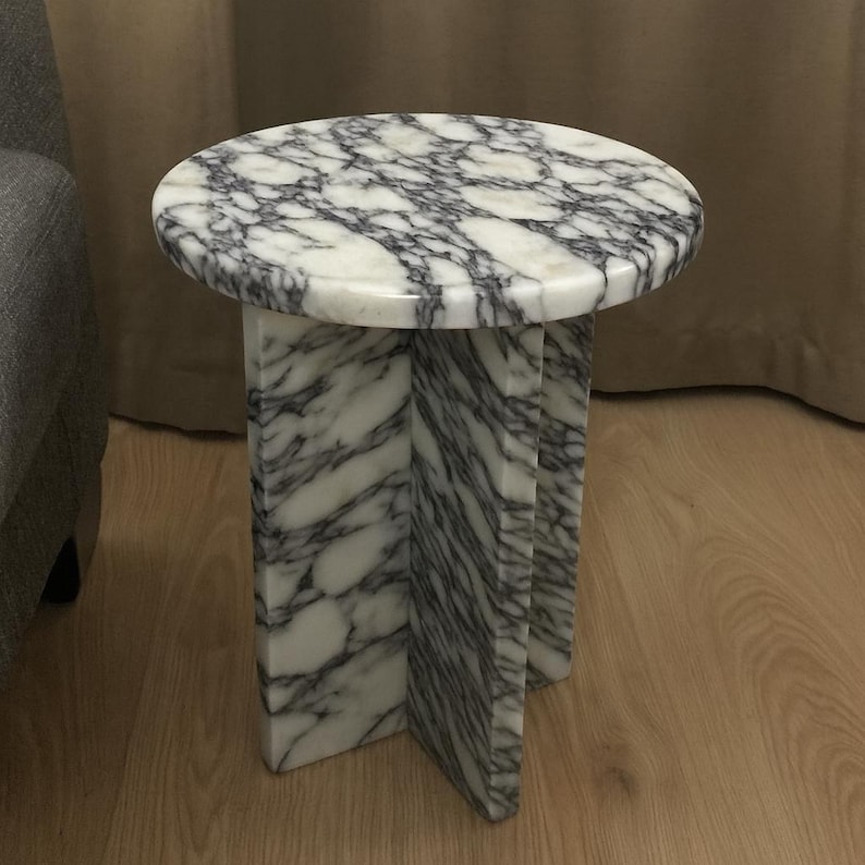May include: A small, round side table with a white marble top and a gray veined pattern. The base is constructed of three intersecting marble slabs. The table is approximately 18 inches in diameter and 18 inches tall.