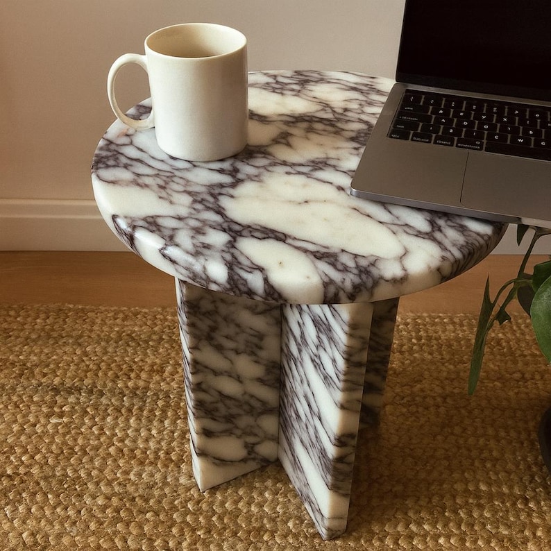 May include: A round marble side table with a white and purple veined pattern. A white mug sits on the table's surface, and a laptop is partially visible. The table has a geometric base and is placed on a woven rug.