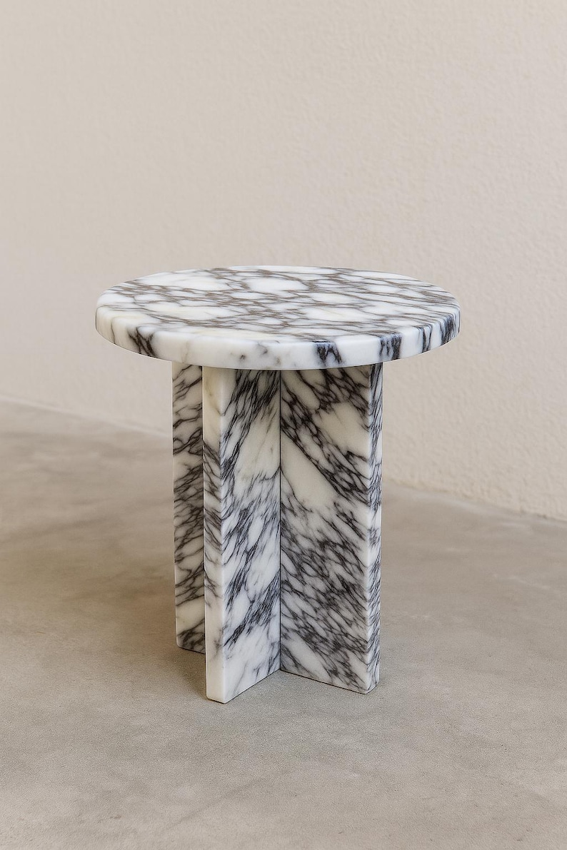 May include: A small, round marble side table with a white and gray veined pattern. The table has a circular top supported by three rectangular legs that intersect at the base. The table is approximately 18 inches in height.