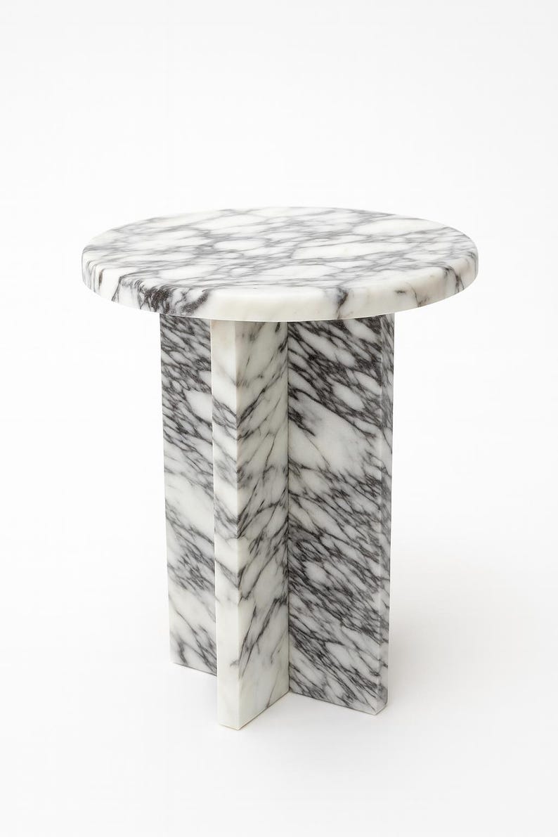 May include: A round marble side table with a white and gray veined pattern. The tabletop is supported by a pedestal base with a cross-shaped design. The table is approximately 20 inches in diameter and 20 inches tall.