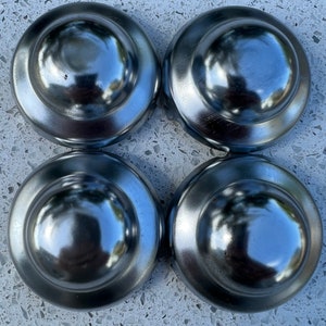 May include: Four shiny, silver-colored hubcaps arranged in a square. Each hubcap has a rounded, domed center and a circular outer rim. The hubcaps reflect the surrounding environment, creating a mirrored effect.