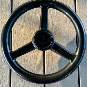 May include: A black, three-spoke steering wheel with a circular center. The wheel is made of a glossy material and has a simple, functional design. The center of the wheel features a small, circular emblem. The wheel is likely for a boat or other watercraft.