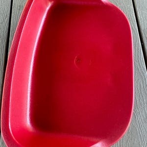 May include: A bright red, plastic tray with a slightly curved shape. The tray has a smooth, glossy surface and a small, circular indentation in the center. The tray is empty and sits on a gray, wooden surface.