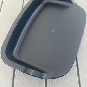 May include: A black rectangular baking dish with rounded corners and a slightly raised edge. The dish has a smooth, matte finish and appears to be made of a durable material. The dish is empty and ready for use.