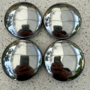 May include: Four shiny, chrome-colored hubcaps arranged in a square. Each hubcap has a rounded, domed shape and reflects the surrounding environment. The hubcaps are likely for a car or truck, adding a classic aesthetic.