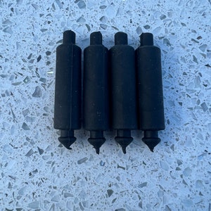 May include: Four black rubber furniture leg tips. Each tip has a cylindrical body with a flared top and a pointed end. The tips are arranged in a row against a speckled, light-colored surface.