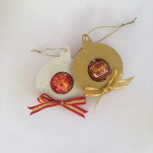 Puede incluir: Dos adornos navide&ntilde;os hechos de papel con brillantina, uno dorado y uno blanco, con una trufa de chocolate Lindt Lindor dentro de cada uno. Ambos adornos tienen una cinta dorada atada alrededor.