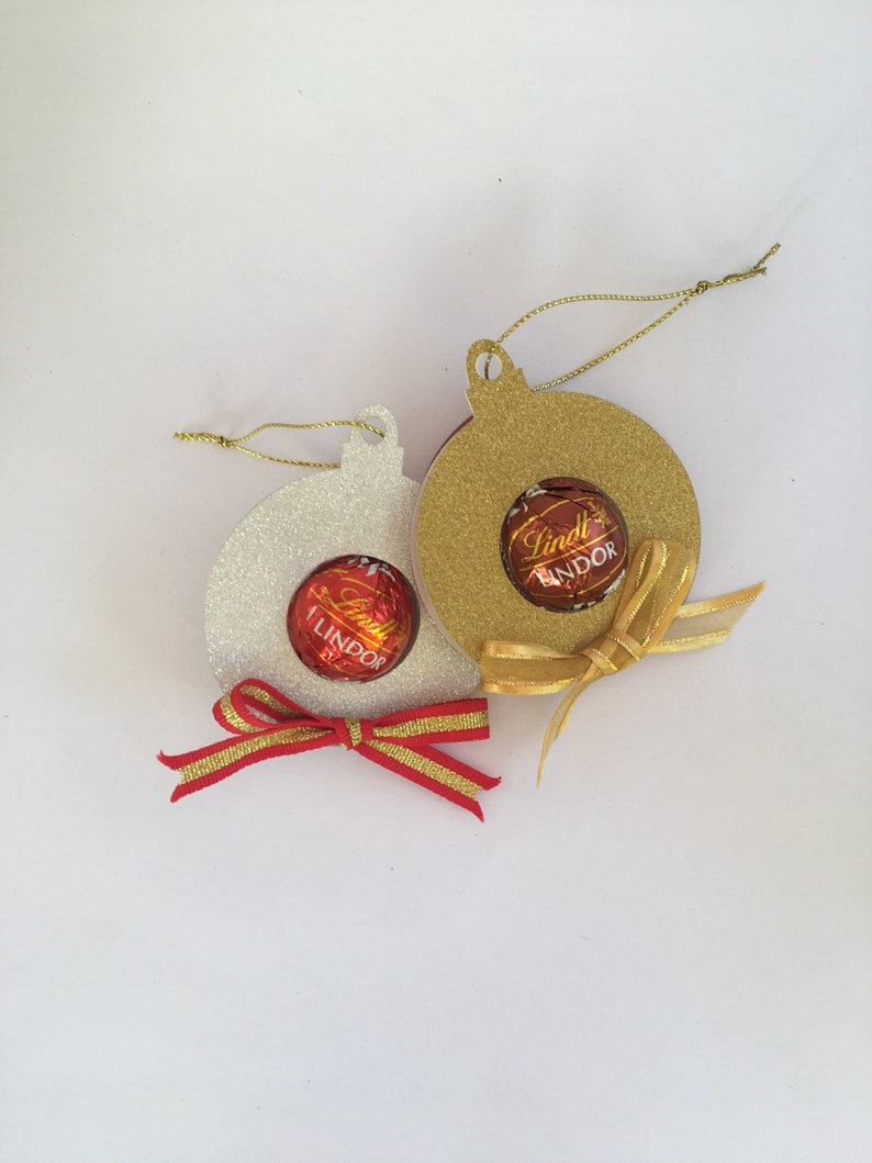Puede incluir: Dos adornos navide&ntilde;os hechos de papel con brillantina, uno dorado y uno blanco, con una trufa de chocolate Lindt Lindor dentro de cada uno. Ambos adornos tienen una cinta dorada atada alrededor.
