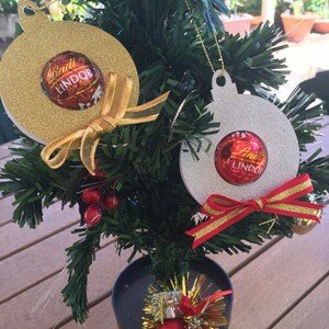 Puede incluir: Dos adornos navide&ntilde;os hechos de cart&oacute;n con purpurina. Los adornos tienen forma de bola y tienen un agujero en la parte superior para colgar. Cada adorno tiene una trufa de chocolate Lindt Lindor en su interior. Un adorno es dorado y el otro es plateado.