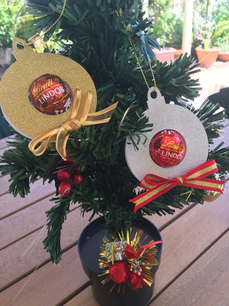 Puede incluir: Dos adornos navide&ntilde;os hechos de cart&oacute;n con purpurina. Los adornos tienen forma de bola y tienen un agujero en la parte superior para colgar. Cada adorno tiene una trufa de chocolate Lindt Lindor en su interior. Un adorno es dorado y el otro es plateado.