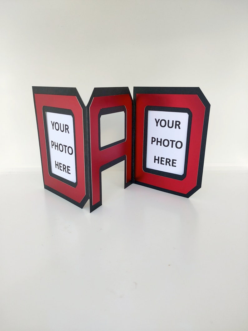 May include: Red and black cardstock photo frame shaped like the word "DAD". The frame has two openings for photos with the text "YOUR PHOTO HERE" printed inside each opening.
