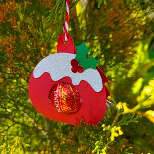May include: Red and white paper Christmas ornament shaped like a pudding with a green holly sprig on top. A chocolate Lindor truffle is visible inside the ornament.