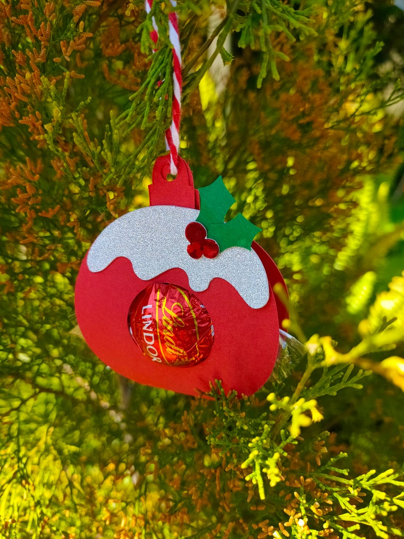 May include: Red and white paper Christmas ornament shaped like a pudding with a green holly sprig on top. A chocolate Lindor truffle is visible inside the ornament.