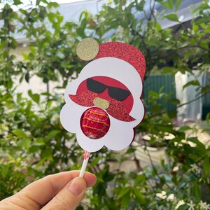 May include: A red and white Santa Claus lollipop holder with a gold glitter hat and sunglasses. The holder has a cut-out for a lollipop.