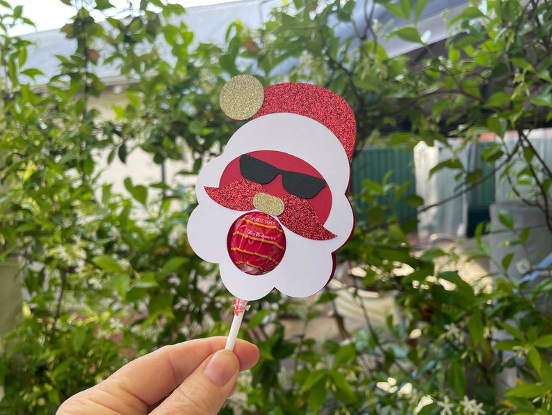 May include: A red and white Santa Claus lollipop holder with a gold glitter hat and sunglasses. The holder has a cut-out for a lollipop.