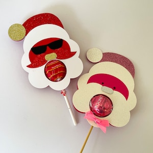 May include: Two red and white Santa Claus lollipop holders with glitter. Each holder has a round opening for a candy. One Santa has sunglasses and a mustache, the other has a pink nose and a bow.