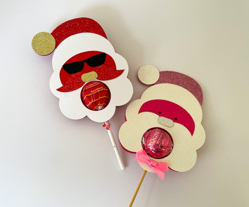 May include: Two red and white Santa Claus lollipop holders with glitter. Each holder has a round opening for a candy. One Santa has sunglasses and a mustache, the other has a pink nose and a bow.