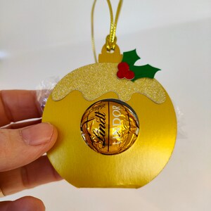 May include: Gold Christmas ornament shaped like a pudding with a Lindt Lindor chocolate inside. The ornament has a green holly leaf with red berries on top.