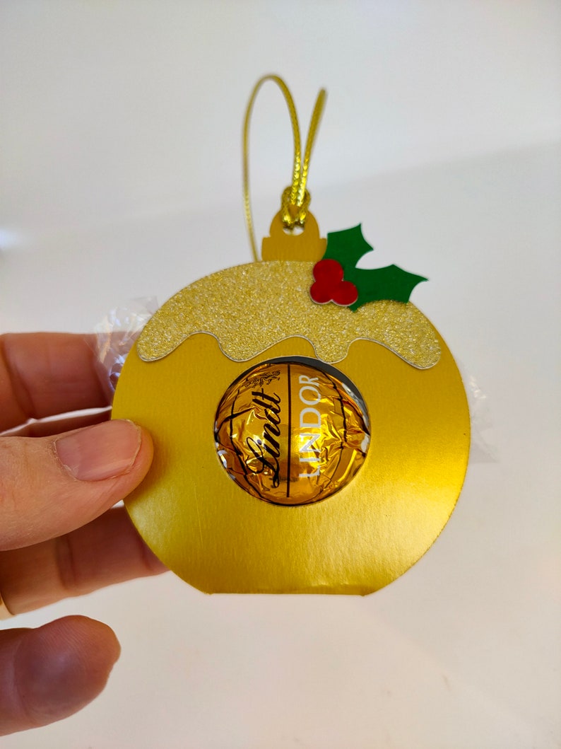 May include: Gold Christmas ornament shaped like a pudding with a Lindt Lindor chocolate inside. The ornament has a green holly leaf with red berries on top.