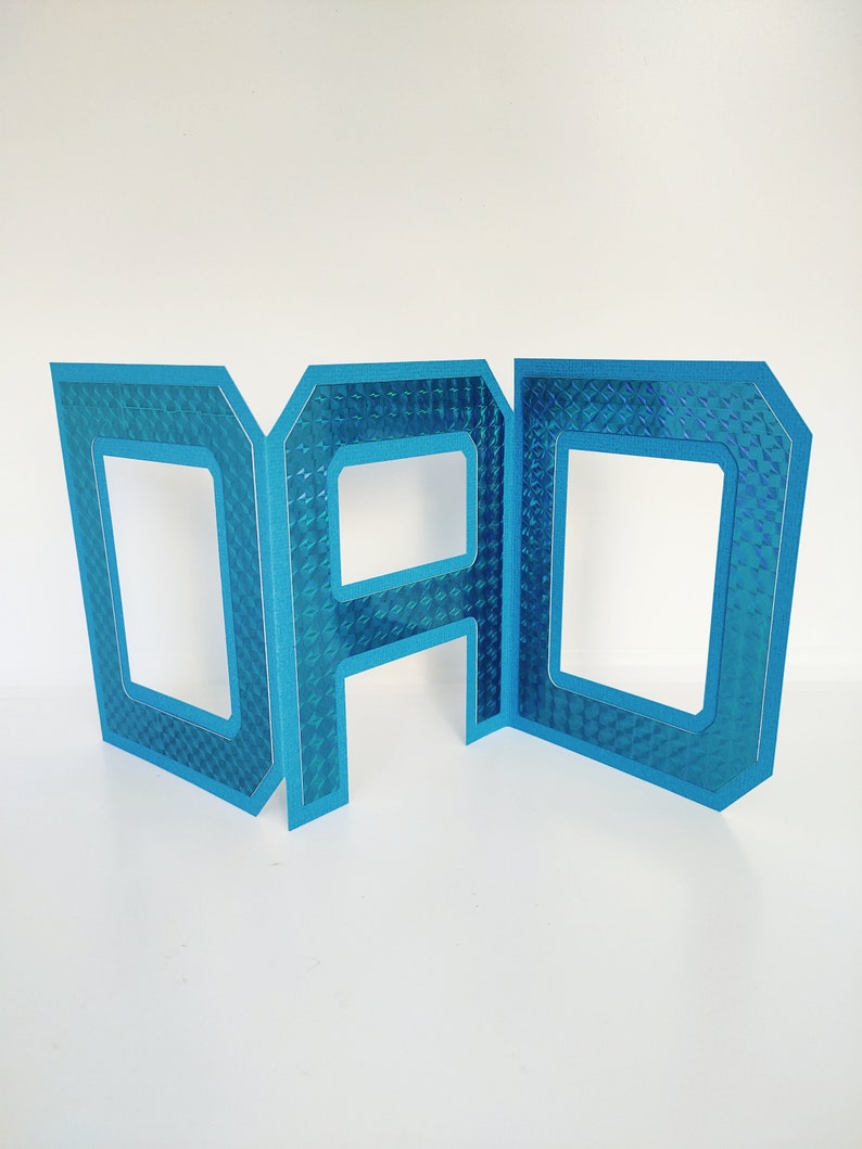 May include: Blue cardboard letters spelling "DAD" with a textured, iridescent finish. Each letter has a cutout in the center.