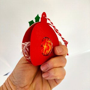 May include: Red paper ornament with a green holly sprig and a white and red string. The ornament is designed to hold a small round candy or treat.